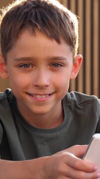 Cheerful boy smiling while holding smartphone outdoors in vertical portrait. Warm sunlight creates a happy childhood lifestyle moment with casual digital interaction in nature. Happy child