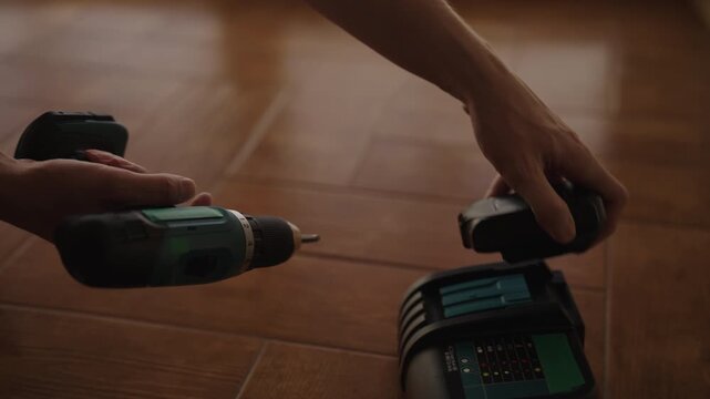Professional worker preparing for job by connecting rechargeable battery to cordless power drill, placing electric screwdriver on wooden floor next to charging station, close-up, slow motion.