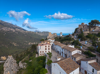 Obraz premium Panoramic view of a picturesque hilltop village in Spain featuring white-washed houses, terracotta rooftops, a medieval castle, and lush green mountains under blue sky