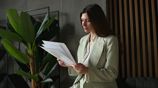 Serious professional woman in a stylish suit sitting in a modern office, carefully reading and analyzing business contracts, reports, or financial paperwork before making a decision