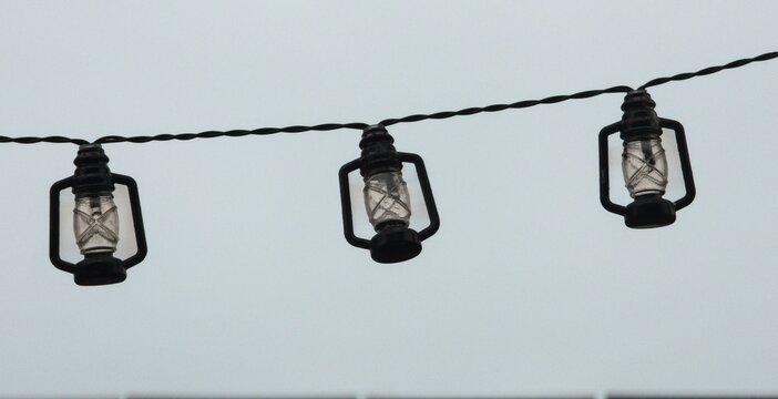 Three identical small black lanterns are strung along a dark cable against a neutral grey sky. The repetitive rhythm of the miniature lamps suggests a cozy outdoor lighting setup for a terrace or even