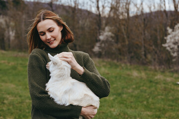 Obraz premium A joyful woman holds a curious goat kid in a sunlit meadow, smiling softly as they share a moment of countryside companionship and rustic lifestyle.