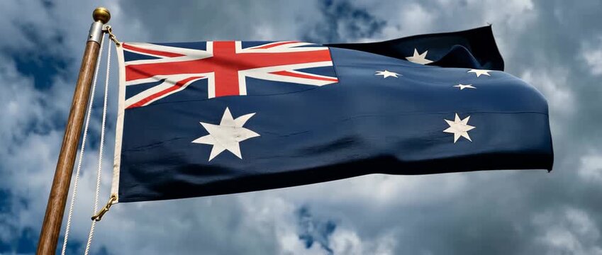 Dramatic close-up of the Australian national flag waving on a wooden flagpole with a finial against a moody cloudy sky, showing Union Jack, Commonwealth Star and Southern Cross.