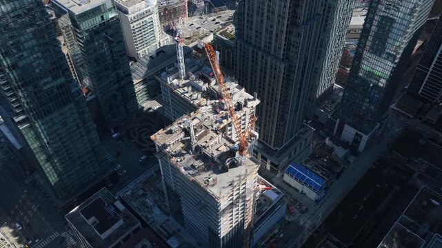 High rise building construction in downtown area with cranes lifting materials over city streets, surrounded by modern office towers and residential skyscrapers