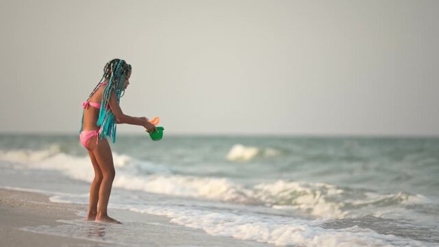 A girl with african braids in a summer costume plays on the beach with shells near the sea with waves under sunny sunset