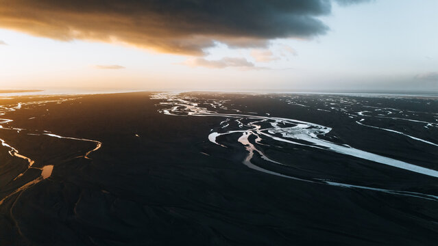 Aerial view of a braided glacial river system flowing through black volcanic sand under a dramatic sunset sky in Skaftarhreppur, Iceland.