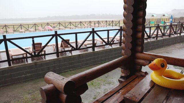 An inflatable duck lies on wooden table in gazebo at recreation center against the backdrop of pool covered with rain