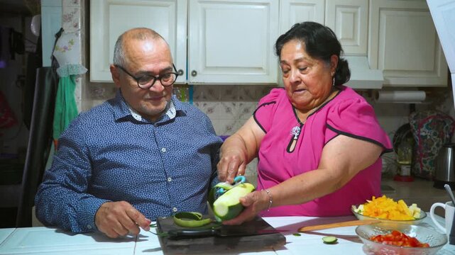 An elderly woman shows her husband how to peel and slice a cucumber on a cutting board
