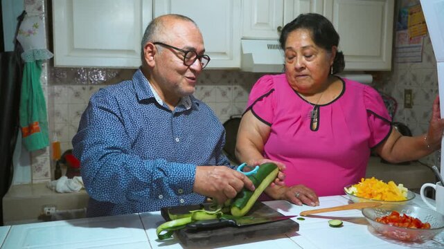 An elderly woman shows her husband how to peel and slice a cucumber on a cutting board
