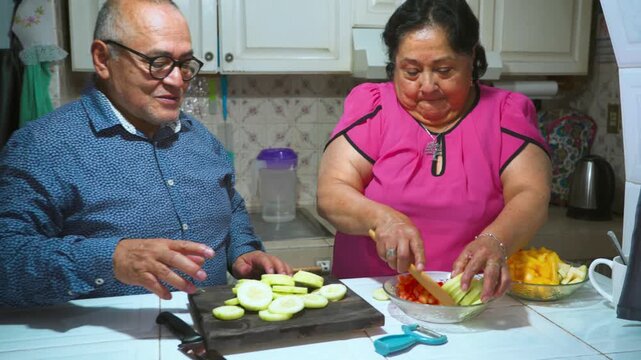 An elderly woman shows her husband how to peel and slice a cucumber on a cutting board