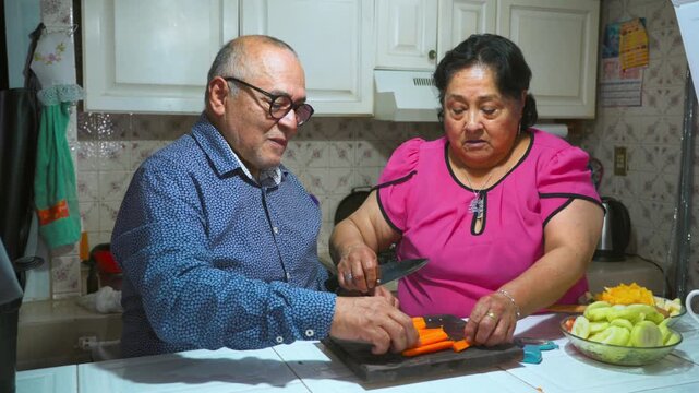 An elderly woman shows her husband how to peel and cut a carrot on a cutting board