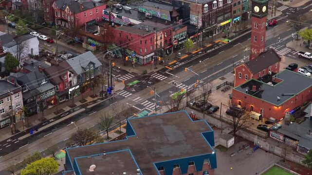 High angle establishing shot of Kensington Market in Toronto on a rainy evening, showing wet asphalt roads, sparse traffic, and glowing storefronts in city's vibrant district