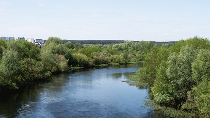 Obraz premium River Flowing Through Lush Green Trees Creating Tranquil Countryside Panorama With Gentle Ripples, Sunlit Sky, Mixed Deciduous Forest Along Meandering Bank, Distant Horizon And Clouds Reflecting