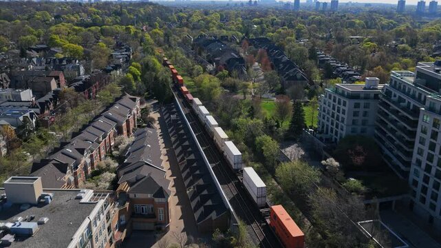 Container freight train running through urban neighborhood with modern apartments, lush green trees, city skyline on sunny day representing industrial logistics