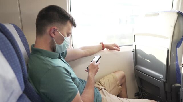 Public transport, man traveling inside train sitting keeping safety distance and mask, sitting using the phone. Male use smart phone inside train car. Traveler wears mask to prevent spread Covid 19.