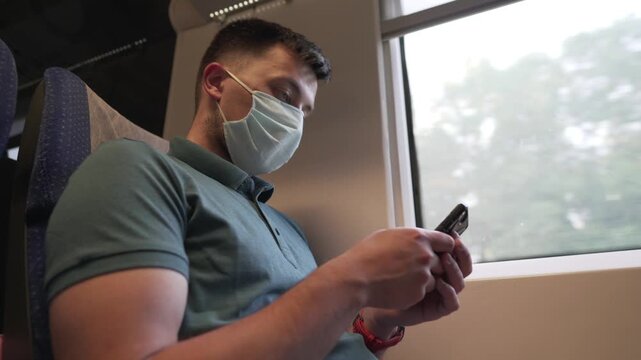 Public transport, man traveling inside train sitting keeping safety distance and mask, sitting using the phone. Male use smart phone inside train car. Traveler wears mask to prevent spread Covid 19.