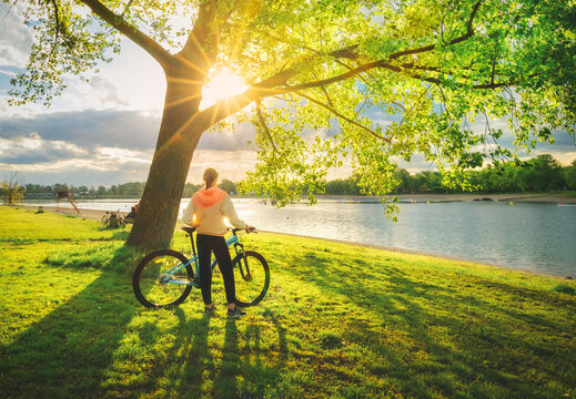 Woman riding a mountain bike near green trees and lake at sunset in spring. Colorful landscape with sporty girl, bicycle, beach, river, green grass, river in park. Summer. Sport and travel. Biking