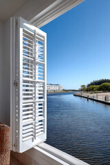 Fototapeta premium White plantation shutters open to tranquil waterfront view, blue sky, calm water, modern architecture, peaceful coastal home