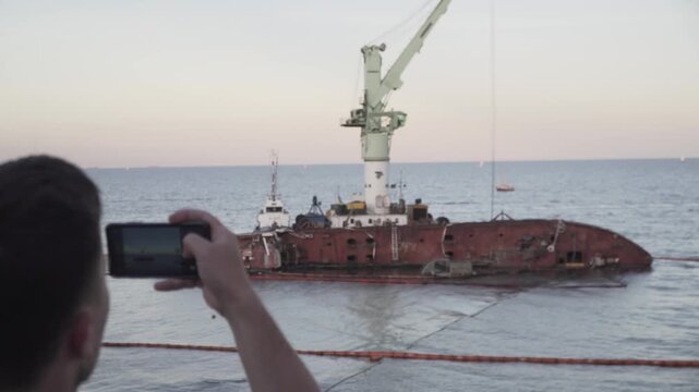 Human journalist makes report on sunken tanker Delfi in Odessa. Male blogger shoots video on phone, publish post about ship wreck in Black Sea. Man video blogger tell subscriptions about boat accident