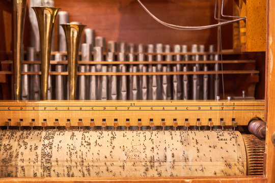 Close-up view of the internal mechanism of a vintage barrel organ or mechanical street organ. The foreground features a rotating wooden cylinder covered with metal pins that encode musical notes,