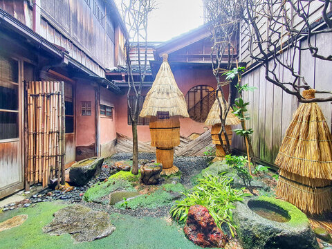 A traditional Japanese courtyard garden (tsuboniwa) situated between historic wooden machiya townhouses, featuring moss-covered ground, large ornamental rocks, and several trees wrapped in straw