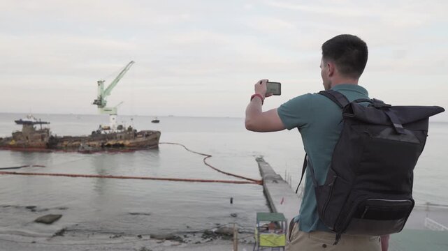 Young traveler blogger takes selfie on phone, makes video broadcast for subscribers on Sea coast about sunken tanker Delf, its rescue using floating crane. Shooting vlog, streaming online podcast.