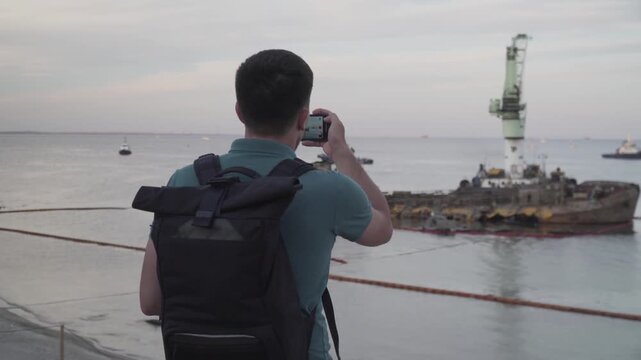 Young traveler blogger takes selfie on phone, makes video broadcast for subscribers on Sea coast about sunken tanker Delf, its rescue using floating crane. Shooting vlog, streaming online podcast.