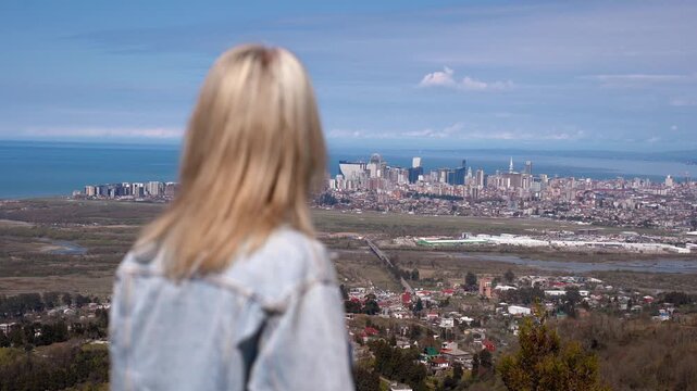 Blonde woman wearing a denim jacket enjoying the beautiful panoramic view of a coastal city with modern buildings and the sea, her hair blowing gently in the wind on a sunny day