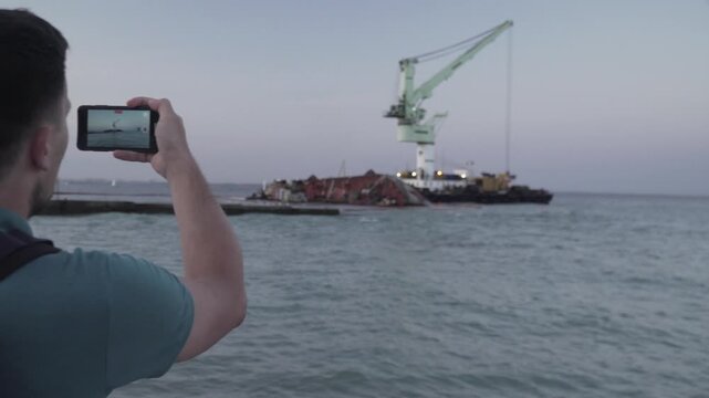 Man blogger records video blog on mobile phone for social media about accident of wreck tanker Delfi with oil in Odessa. Journalist reports on pollution of sea with oil products. Ecology problems.