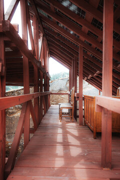 Low view along a wooden gallery under photovoltaic roof panels, with antique chairs, red timber beams, stone walls, and warm rustic sustainable architecture