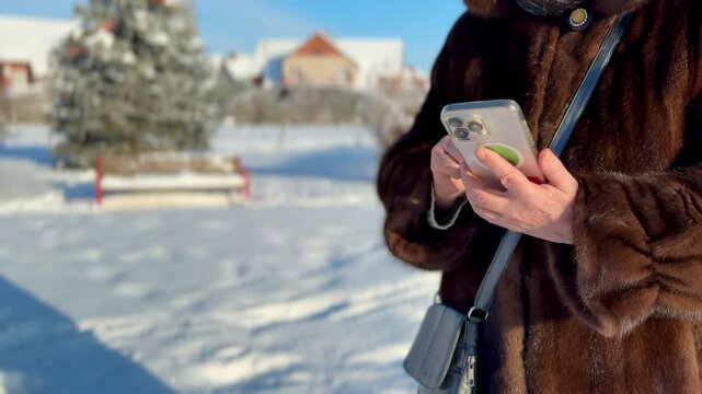 Woman in mink coat texting closeup, hands holding phone in snowy park, severe frost on fur, crossbody bag at side