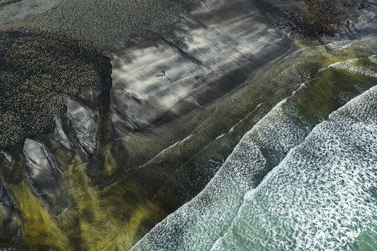 Aerial view of Talisker Bay Beach with dark sand and white waves crashing on the shore as two people walk along the coast Isle of Skye, Scotland, United Kingdom.