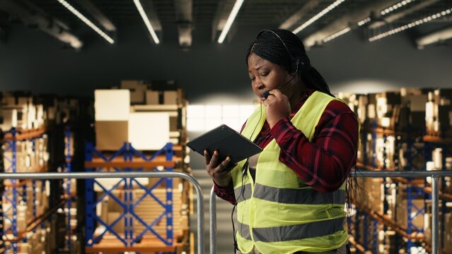 African american woman, customer service complaints, phone call on headset, verifying package tracking info and express delivery updates. Woman operator working on shipment in warehouse.