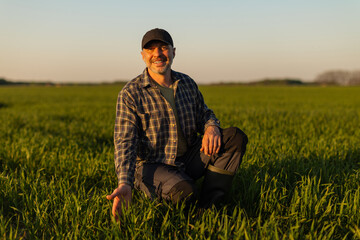 Happy man farmer kneeling in green field examining crops, smiling at camera, working in rural farming area. Successful farm production concept © Home-stock