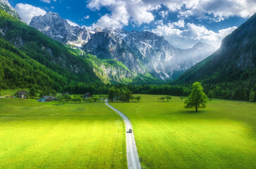 Naklejka premium Aerial view of road in alpine mountains, green meadows, trees in summer. Top view of rural road. Colorful landscape with road, car, rocks, field, grass, sky, sunbeam, clouds. Logar valley, Slovenia