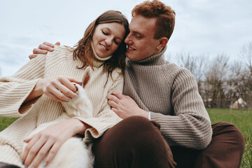 Obraz premium Smiling couple in chunky sweaters outdoors, caressing a fluffy white dog, capturing a cozy moment of love and companionship. Natural light, soft shadows, rural backdrop, relaxed mood, wellness and