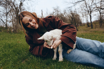 Obraz premium Cheerful woman outdoors, lying on grass with a gentle goat, kid nearby, warm smile, relaxed pose, soft natural light, casual jacket, autumnal park setting, close bond, nature atmosphere