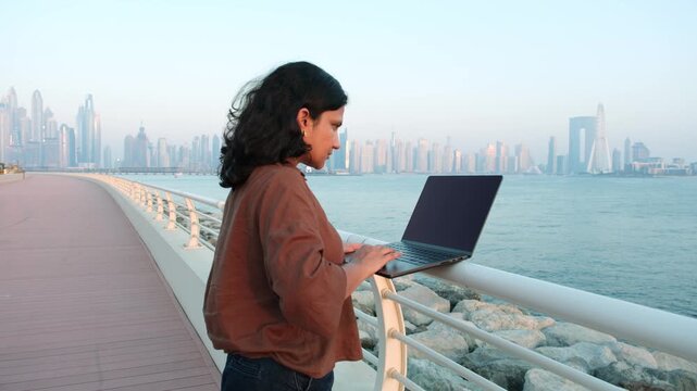 Modern professional woman using laptop by Dubai waterfront during sunset remote work footage