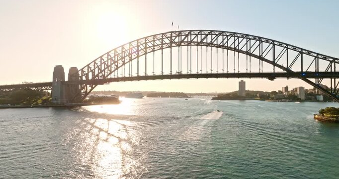 Sydney Harbour Bridge arching over sunlit harbour waters at sunset, motorboats and ferries leaving wakes across golden reflections, iconic city skyline, drone flight aerial view. Explore Australia