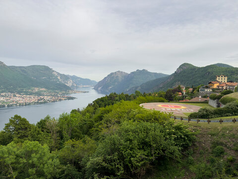 Lake Como is stretching through a mountainous landscape, surrounded by lush green trees and a charming village featuring a helipad with civenna written on it