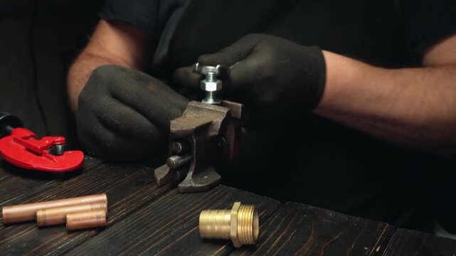Male technician in black gloves tightens a metal bolt using a vise on a wooden workbench with various plumbing components nearby