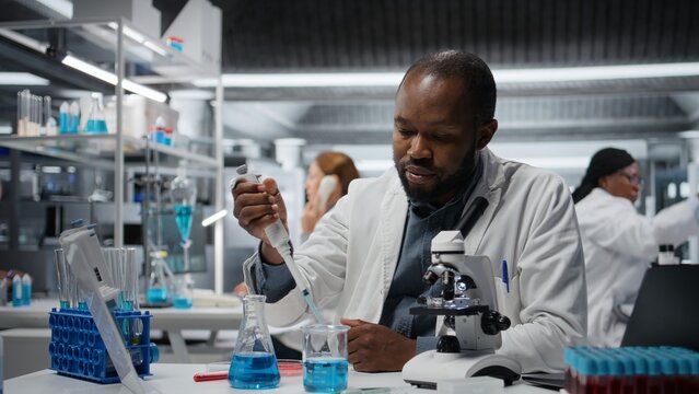 Analytical chemist pipetting blue liquid into test tube to analyze it. African american man transferring chemical sample in glass flask to prepare for spectroscopic analysis, camera A