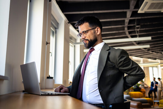 Businessman experiencing back pain while working on laptop in modern office, feeling discomfort and stress at work.