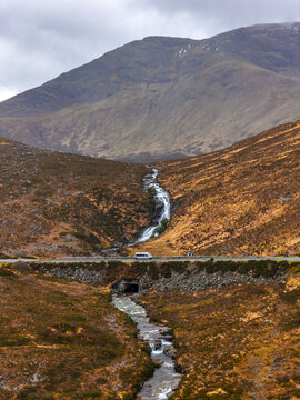 Aerial view of Eas a' Bhradain waterfall cascading down a rugged hillside towards a road with a parked van in Isle of Skye, Scotland, United Kingdom.