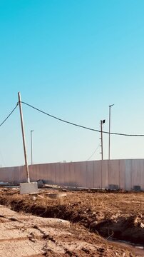 Construction site stainless steel fence against clear blue sky with lighting poles, utility wires, concrete bases and rough gravel