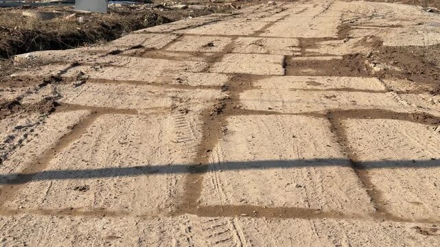 Temporary concrete slab road covered mud, sunlit construction corridor with deep tire tracks, sand packed joints between slabs