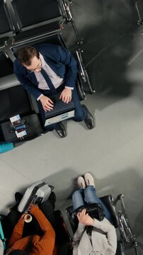Vertical video Multiethnic travelers in airport hub going to boarding gate, carrying wheeled luggage. Diverse people in airport lounge standing up from metal benches, preparing for boarding call