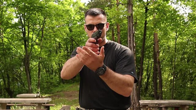 Focused man in sunglasses raising and aiming a black airsoft pistol in a forest clearing, preparing for target shooting practice outdoors with tactical stance and natural green background.