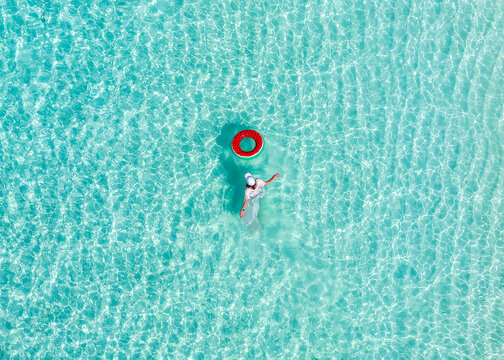Aerial view of a woman wading in crystal clear turquoise water next to a red inflatable ring in Vashafaru, Haa Alif Atoll, Maldives.