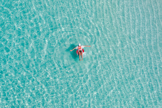 Aerial view of a person wearing a white hat and shirt floating on a red inflatable ring in crystal clear turquoise water with sandy ripples in Vashafaru, Haa Alif Atoll, Maldives.